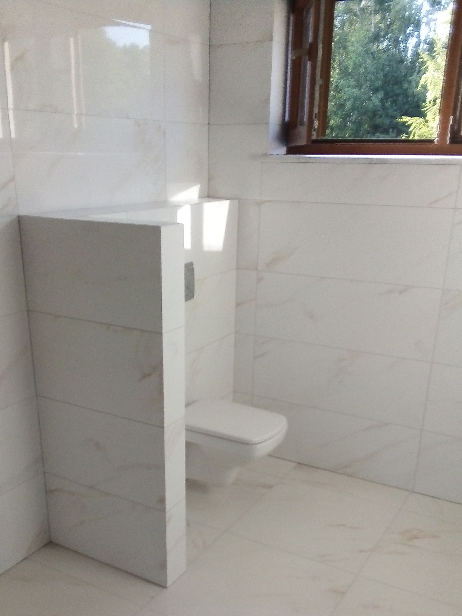 Wykończona łazienka z podwieszaną toaletą w zabudowie, białe płytki z delikatnym marmurkowym wzorem na ścianach i podłodze, drewniane okno z widokiem na zieleń na zewnątrz.