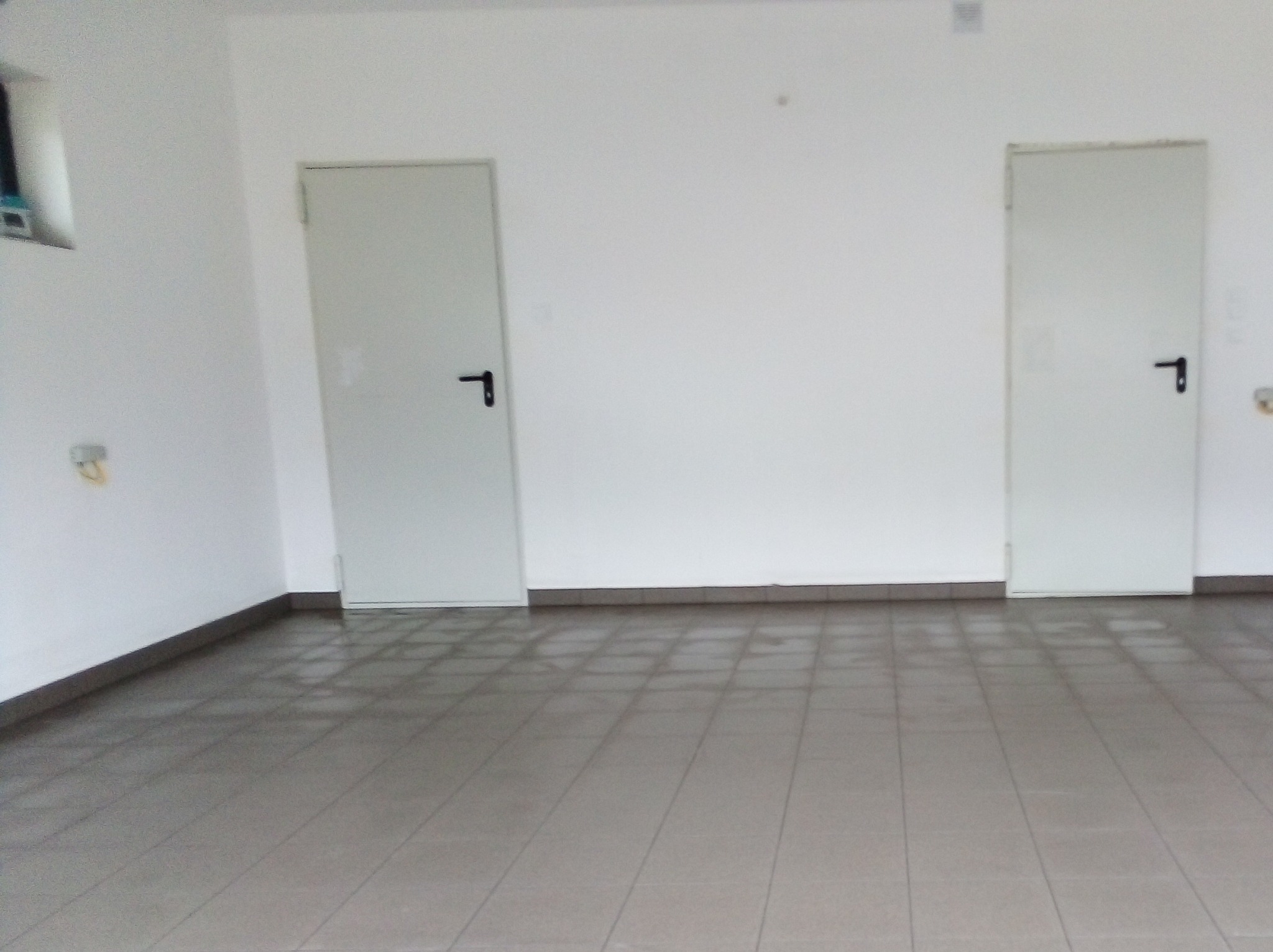 Puste pomieszczenie z dwiema białymi drzwiami z czarnymi klamkami i szarą, błyszczącą podłogą z płytek w kratkę, widoczne detale instalacji elektrycznej na ścianie.
