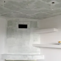 Wnętrze w trakcie przygotowania do malowania: surowy sufit z widocznymi przewodami oświetleniowymi, ściany z widocznymi śladami szpachlowania, kominek z szarego kamienia i białe półki wiszące.