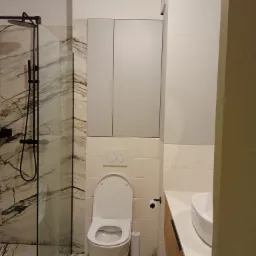 Nowoczesna łazienka z kabiną prysznicową typu walk-in z marmurowymi płytkami, wiszącą toaletą z podtynkową spłuczką i szarą szafką nad nią oraz umywalką nablatową na drewnianej szafce.
