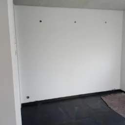 Wnętrze pokoju w trakcie remontu: białe ściany z widocznymi otworami po gniazdkach i punktach świetlnych, czarna podłoga zabezpieczona matą ochronną.