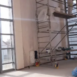 Wnętrze w trakcie prac wykończeniowych: pracownik na rusztowaniu, ściany przygotowane do malowania, duże okna, widoczny grzejnik i narzędzia malarskie.