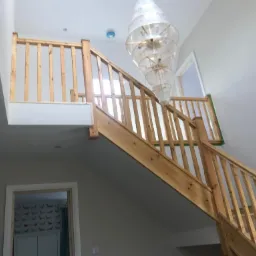 Wnętrze domu w trakcie prac wykończeniowych: drewniane schody z balustradą, widoczna lampa wisząca i fragment ściany z tapetą w tle.