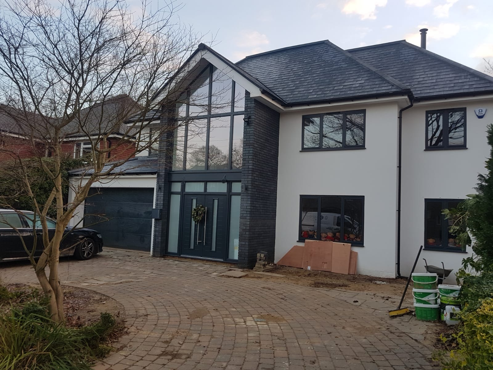 Nowoczesny dom z szarym dachem, białymi ścianami i dużym, przeszklonym wejściem, z widocznymi materiałami budowlanymi i narzędziami na podjeździe.