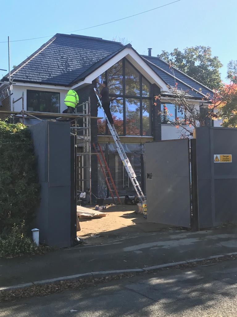 Dwóch pracowników na rusztowaniu i drabinie montuje okna w nowoczesnym domu z ciemną elewacją, widoczne narzędzia i materiały budowlane.