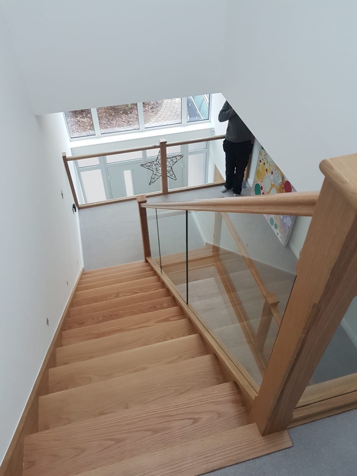 Widok z góry na drewniane schody z balustradą ze szkła i drewna w nowoczesnym domu; w tle widoczny fragment osoby i dekoracja w kształcie gwiazdy.