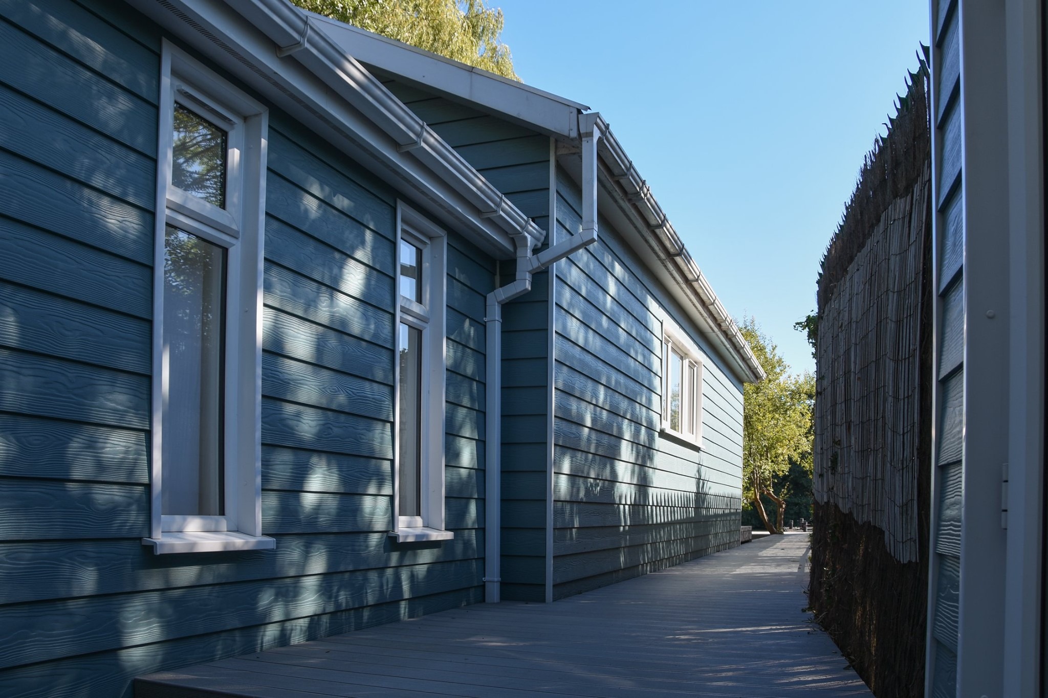 Boczny widok domu z niebieską elewacją z desek, białymi oknami i szarym dachem, z widocznym drewnianym tarasem i fragmentem maty trzcinowej na granicy działki.