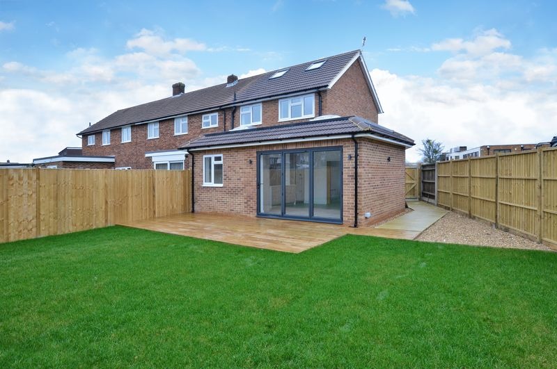 Rozbudowany dom z cegły z przeszklonymi drzwiami harmonijkowymi wychodzącymi na drewniany taras i zadbany trawnik, widok z ogrodu.