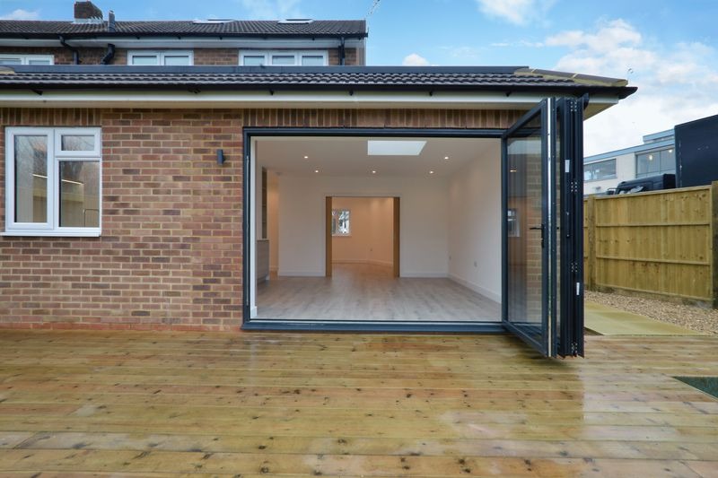 Rozbudowa domu z cegły: widok z zewnątrz na otwarte, składane drzwi przesuwne prowadzące do jasnego, wykończonego wnętrza z drewnianą podłogą i oknem dachowym, na zewnątrz drewniany taras.