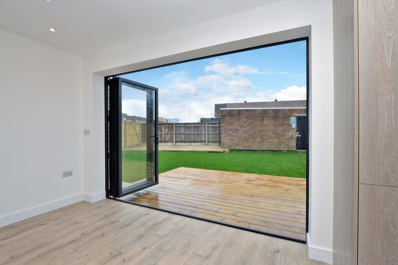 Nowoczesne, składane drzwi balkonowe z czarną ramą, otwarte na drewniany taras i zielony trawnik, widok z wnętrza jasnego pomieszczenia z panelami podłogowymi.