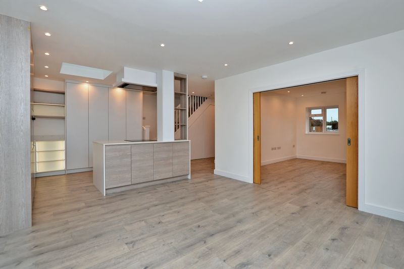 Jasne, przestronne wnętrze domu po remoncie z drewnianą podłogą, zabudową kuchenną w odcieniach szarości i bieli, oświetleniem punktowym i przesuwnymi drzwiami do drugiego pomieszczenia z oknem.