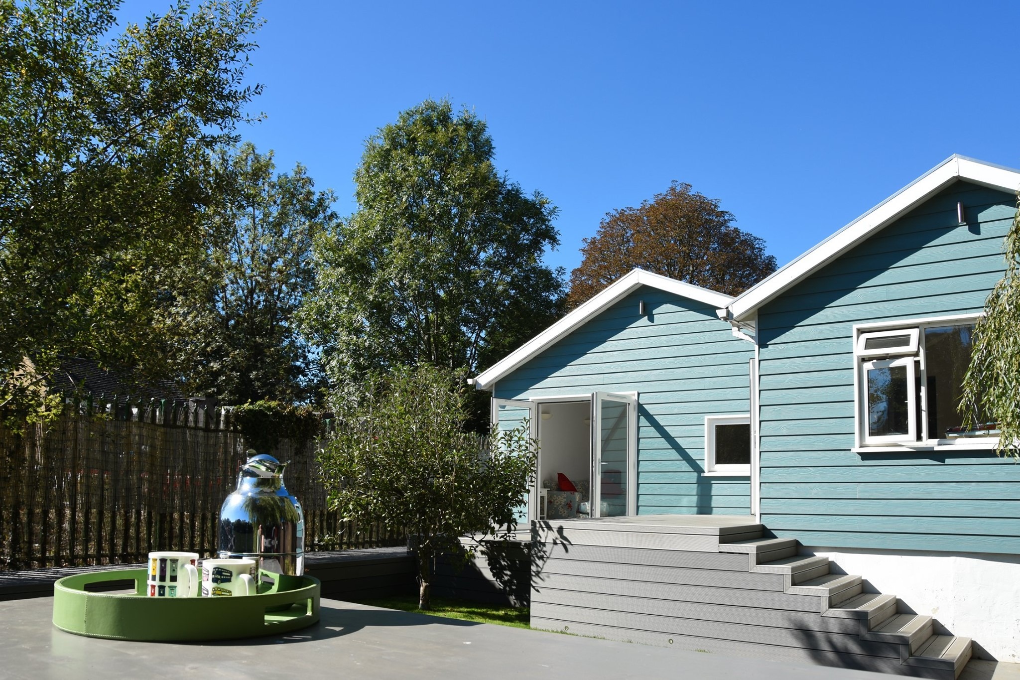 Niebieski dom z drewnianą elewacją i szarym tarasem z kompozytu, widok z zewnątrz w słoneczny dzień, na pierwszym planie stolik z zieloną tacą, dzbankiem i kubkami.