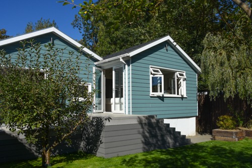 Niebieski dom z drewnianą elewacją, białymi oknami i drzwiami balkonowymi, otoczony zielenią ogrodu. Widoczne schody prowadzące do tarasu z desek kompozytowych.