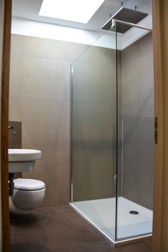 Nowoczesna łazienka z kabiną prysznicową typu walk-in, umywalką wiszącą i brązowymi płytkami podłogowymi, doświetlona światłem dziennym z okna dachowego.