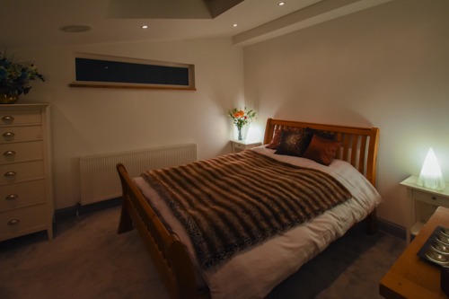 Jasne wnętrze sypialni z drewnianym łóżkiem przykrytym brązowym pledem, biała komoda z kwiatami, nocne stoliki z lampkami i małe okno pod sufitem.