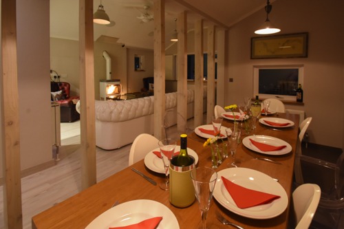 Ustawiony stół w jasnym wnętrzu domu z drewnianą przegrodą i kominkiem w tle, widoczne nakrycia z czerwonymi serwetkami, butelka wina i kwiaty.