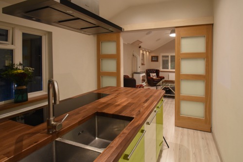 Nowoczesna kuchnia z drewnianym blatem, stalowym zlewem i przesuwnymi drzwiami z matowego szkła prowadzącymi do salonu.