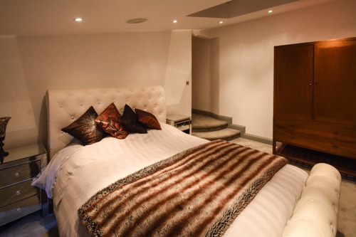 Sypialnia z łóżkiem z pikowanym zagłówkiem, lustrzanymi szafkami nocnymi, futrzanym pledem w brązowe pasy i drewnianą szafą w tle, z widocznymi schodami prowadzącymi do innego pomieszczenia.