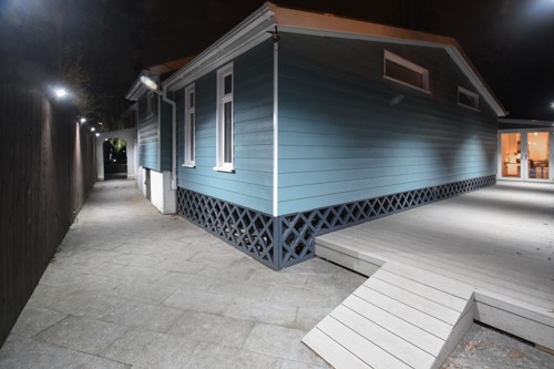 Nocne ujęcie bocznej ściany domu z elewacją w odcieniach błękitu, z ozdobnym ażurowym cokołem i drewnianym tarasem z rampą, oświetlonego zewnętrznymi lampami.