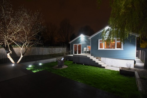 Nocne ujęcie domu z niebieską elewacją, oświetlonego punktowymi lampami w ogrodzie, z widocznymi schodami prowadzącymi do drzwi tarasowych i drzewem z podświetlonym pniem.