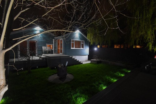 Nocne ujęcie domu z drewnianą elewacją i oświetlonym ogrodem, widoczne drzewo z gołymi gałęziami na pierwszym planie, oświetlenie elewacyjne i ogrodowe.