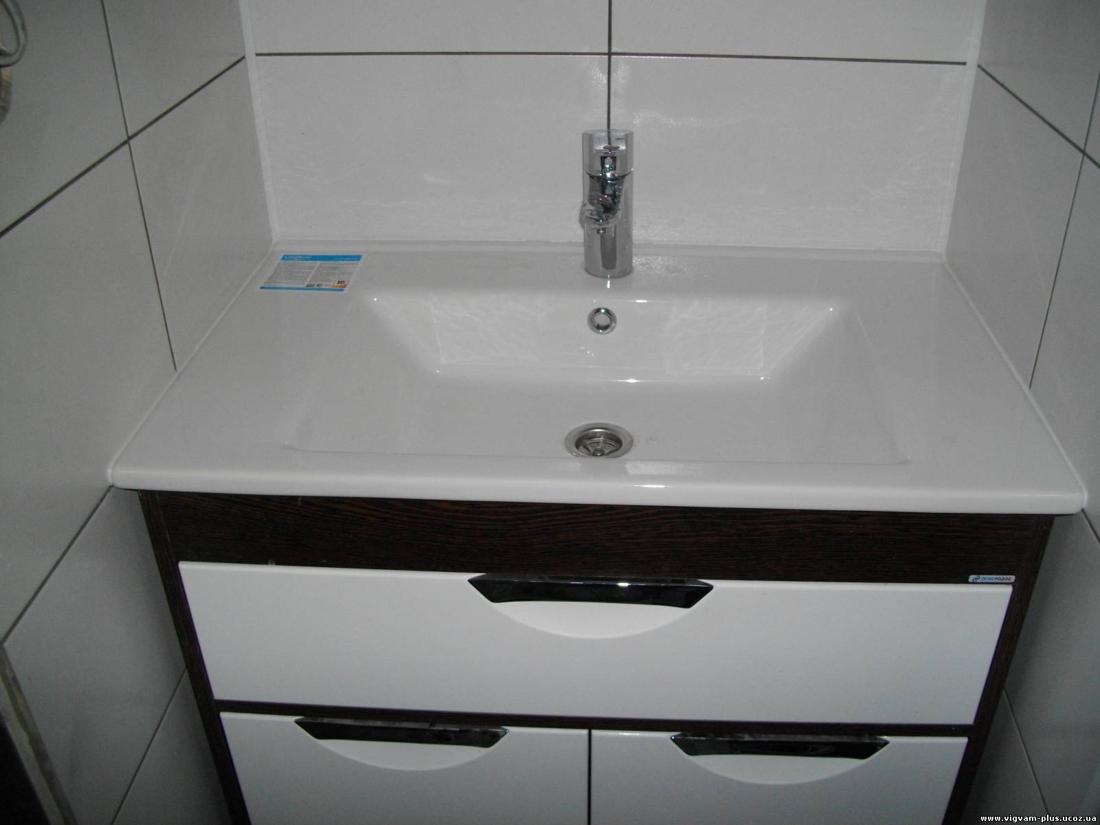 Biała umywalka z chromowaną baterią i odpływem, osadzona na szafce z ciemnobrązowym blatem i białymi, zaokrąglonymi szufladami z czarnymi uchwytami, na tle białych płytek ściennych z ciemnoszarą fugą.
