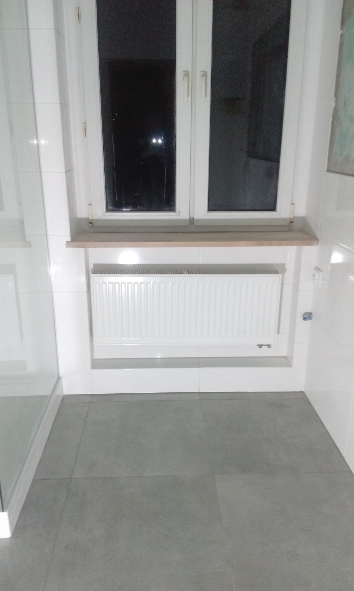 Biała łazienka z grzejnikiem pod oknem, szare płytki podłogowe i fragment kabiny prysznicowej.
