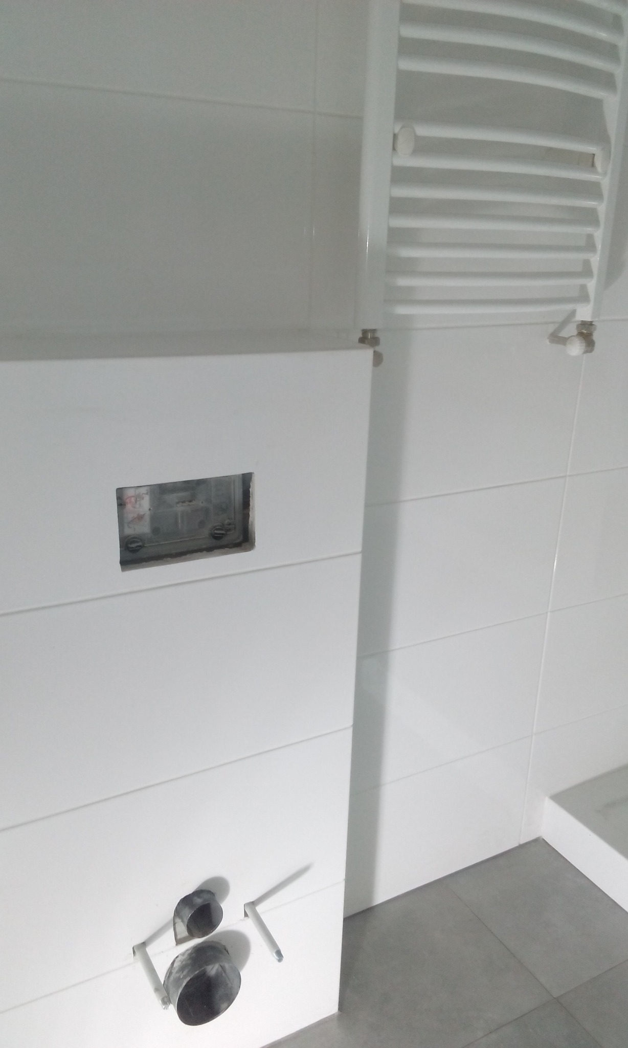 Biała, częściowo wykończona łazienka z widoczną instalacją podtynkową, grzejnikiem drabinkowym i szarymi płytkami podłogowymi.