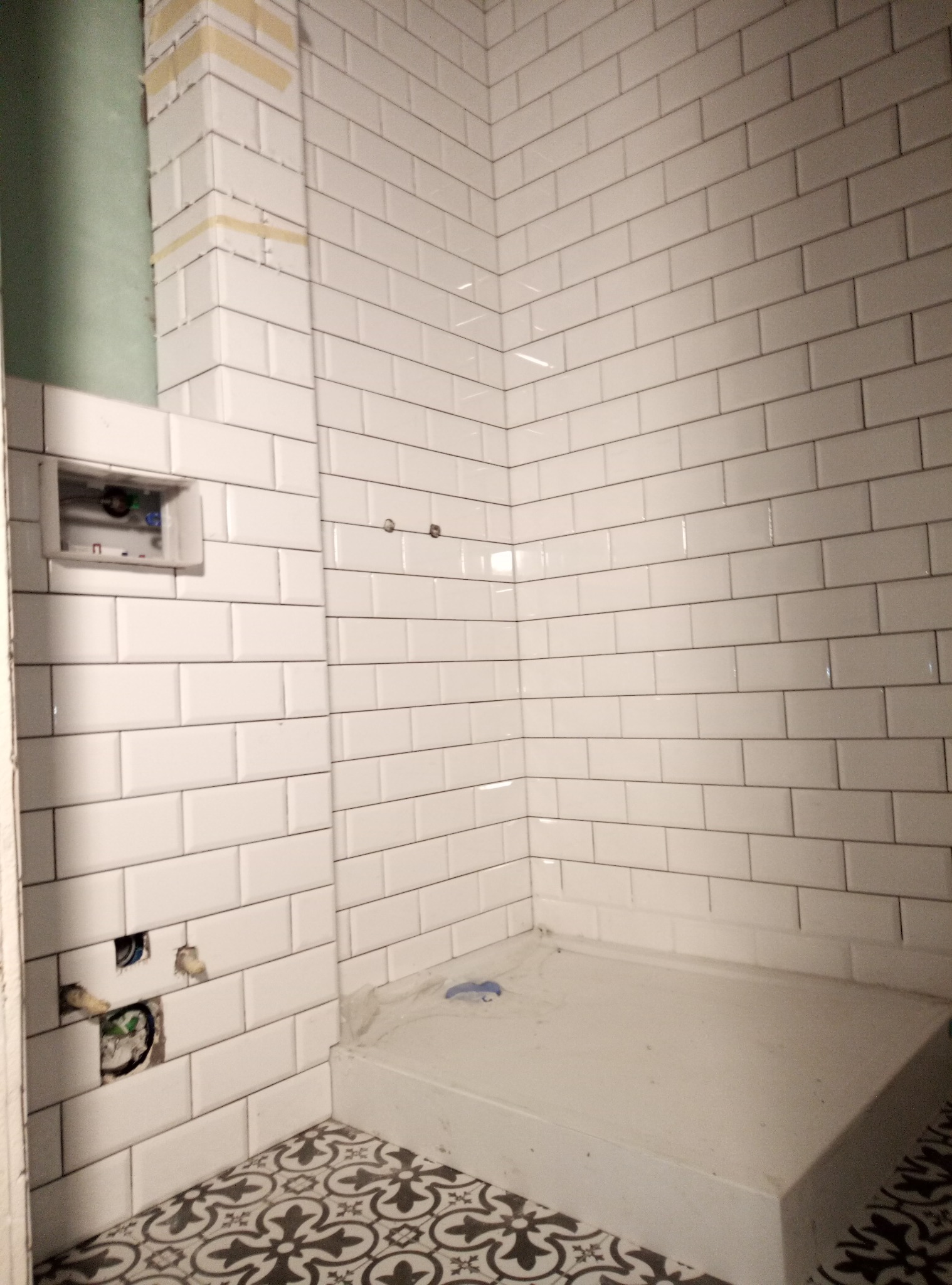 Świeżo ułożone białe, prostokątne płytki w łazience, z widocznymi otworami na instalacje i fragmentem dekoracyjnej podłogi w stylu vintage.