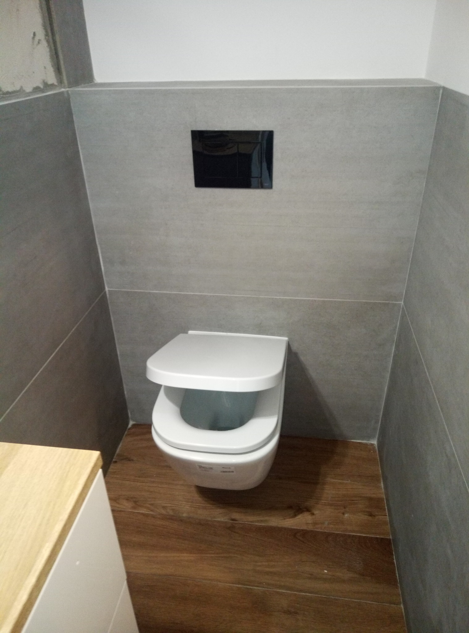 Nowoczesna toaleta podwieszana z szarymi, matowymi płytkami na ścianach i drewnopodobną podłogą, widoczny fragment szafki pod umywalkę z blatem w kolorze jasnego drewna.