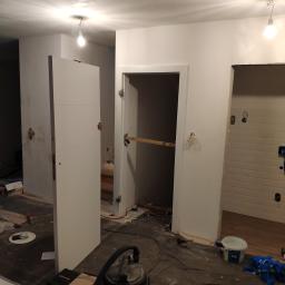 Rubin Technology - Montaż białych drzwi wewnętrznych z trzema poziomymi frezowaniami w nowo wykończonym wnętrzu, widoczne ślady prac remontowych i odkurzacz przemysłowy.