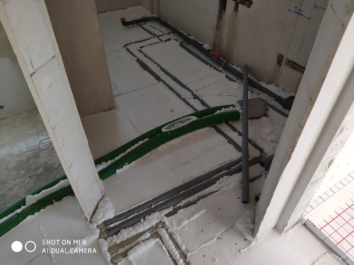 Przygotowanie podłoża pod ogrzewanie podłogowe: ułożona izolacja ze styropianu z wyciętymi kanałami na rury, widoczne rury instalacji sanitarnej i przewody elektryczne w karbowanej osłonie.