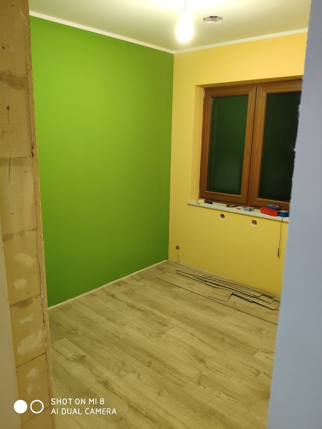 Pokój po malowaniu ścian na zielono i żółto, widoczne przygotowane panele podłogowe, gniazdka elektryczne bez osłon i drewniane okno z narzędziami na parapecie.