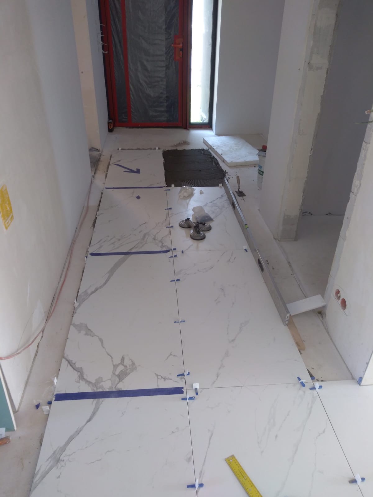 Układanie białych płytek imitujących marmur na podłodze w korytarzu, widoczne przyssawki do przenoszenia płytek, poziomica i niebieska taśma maskująca.