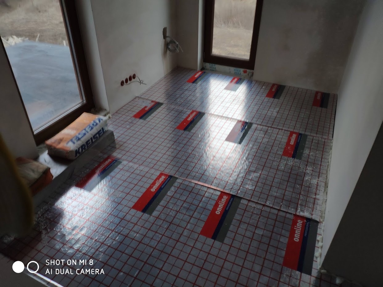 Przygotowanie podłoża pod ogrzewanie podłogowe w surowym wnętrzu. Widoczne maty grzewcze z czerwoną kratką i napisem 'onnline professional' rozłożone na podłodze, obok okna leży worek zaprawy Kreisel.