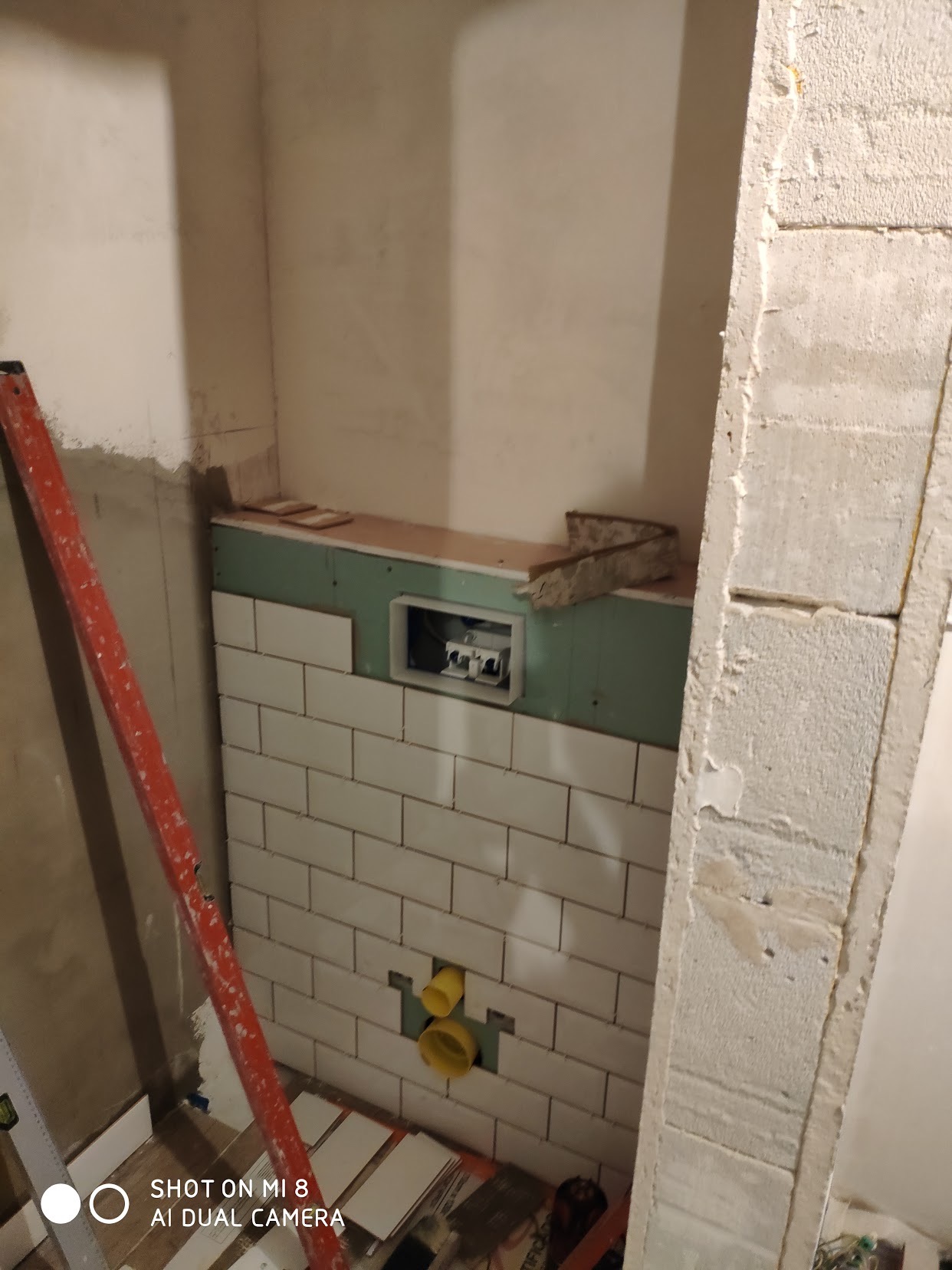 Ściana w trakcie wykańczania białymi, prostokątnymi płytkami, widoczne żółte rury instalacyjne, zielona płyta gipsowo-kartonowa, poziomica i fragment murowanej ściany w surowym stanie.