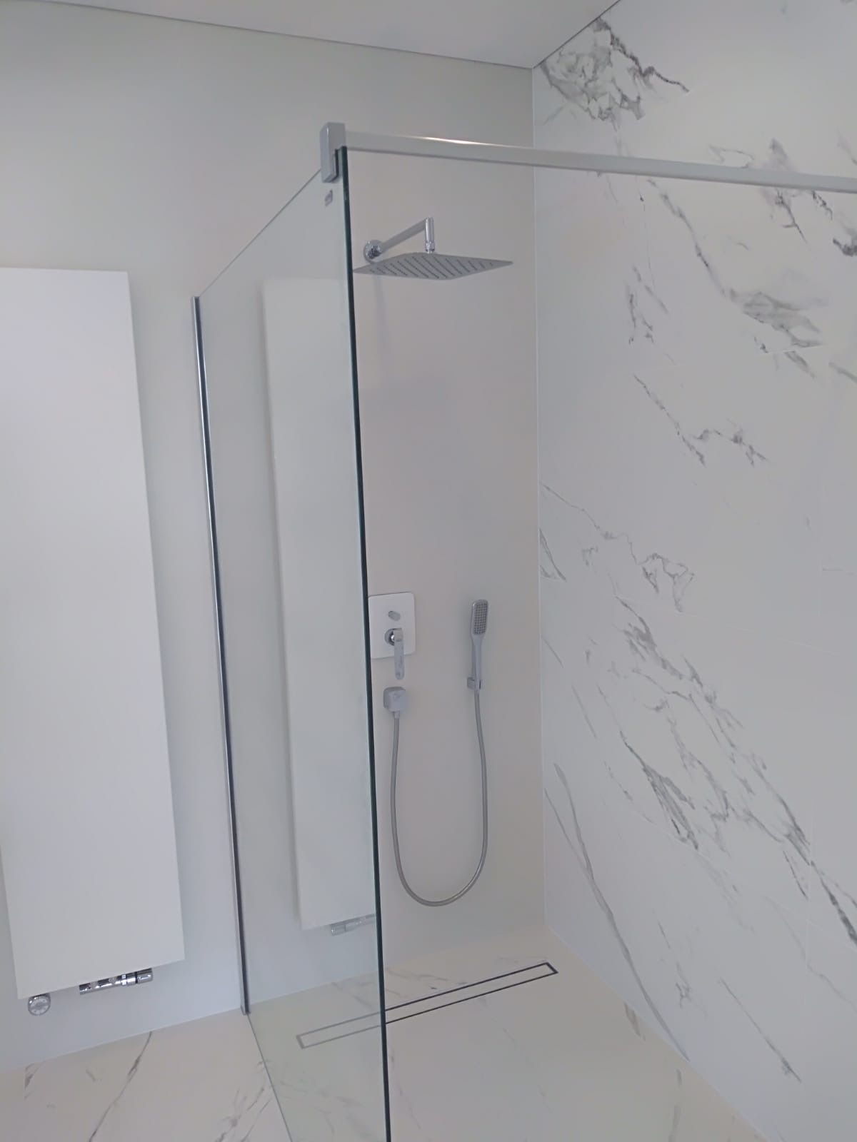 Nowoczesna kabina prysznicowa walk-in z deszczownicą, panelem sterowania i odpływem liniowym w podłodze, wykończona białymi płytami imitującymi marmur.