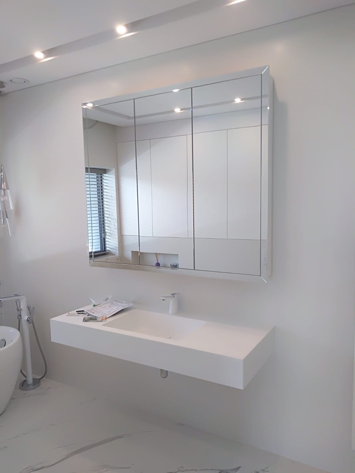 Minimalistyczna łazienka w bieli: podwieszana szafka z umywalką, lustro z szafkami, marmurowa podłoga z szarymi żyłami, widoczne narzędzia i instrukcja montażu na blacie.