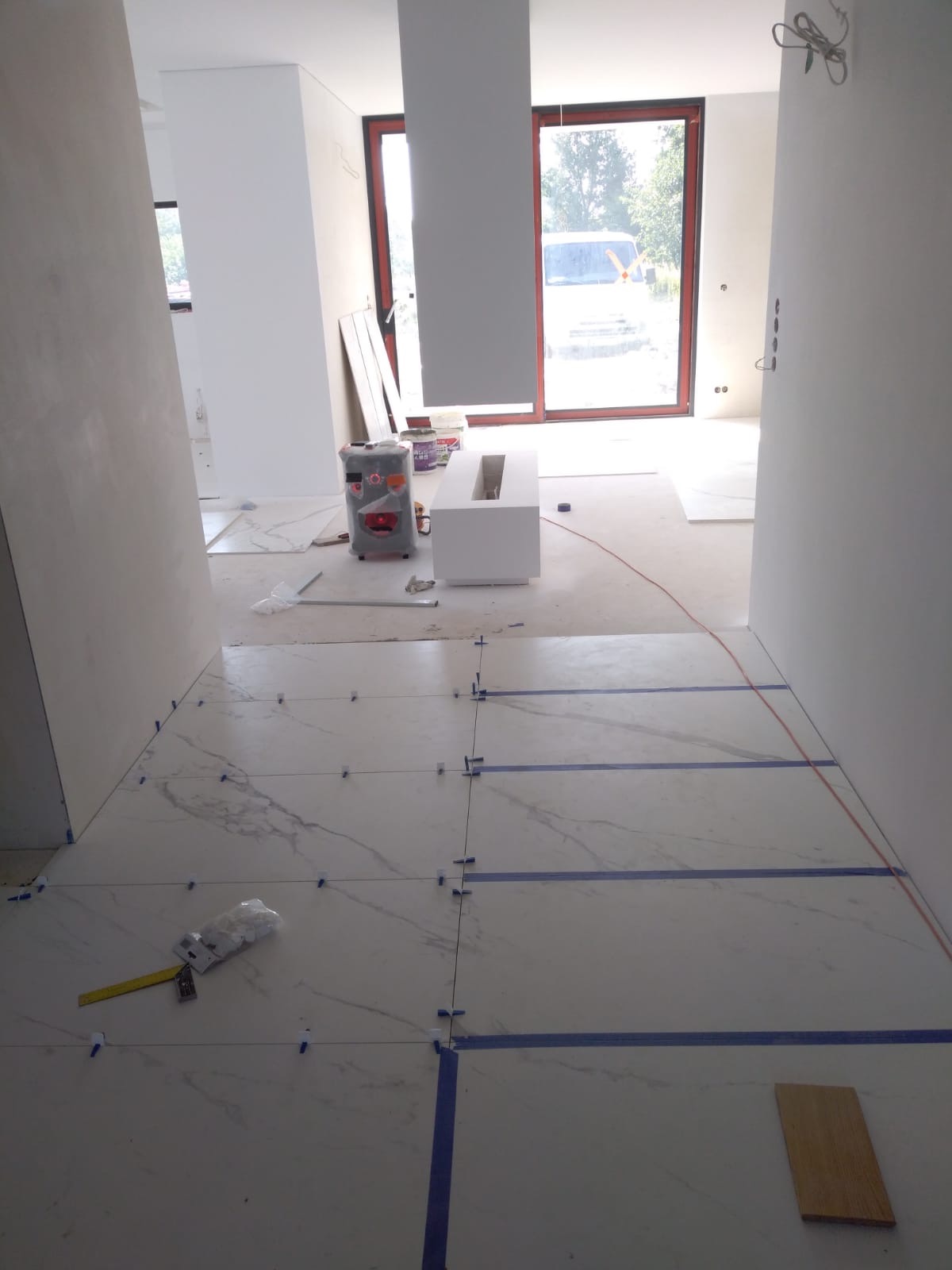 Wnętrze w trakcie układania marmurowej podłogi: widoczne płytki z niebieską taśmą wyznaczającą linie, krzyżyki dystansowe, poziomica i paczka z zapasem krzyżyków.