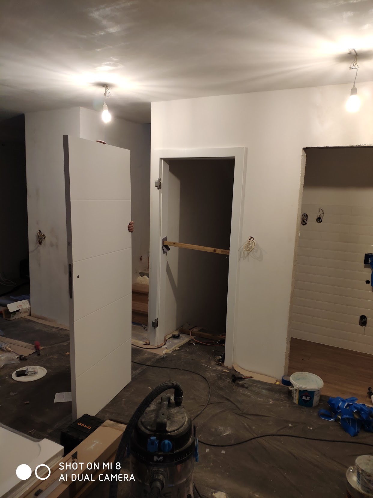Montaż białych drzwi wewnętrznych z trzema poziomymi frezowaniami w nowo wykończonym wnętrzu, widoczne ślady prac remontowych i odkurzacz przemysłowy.