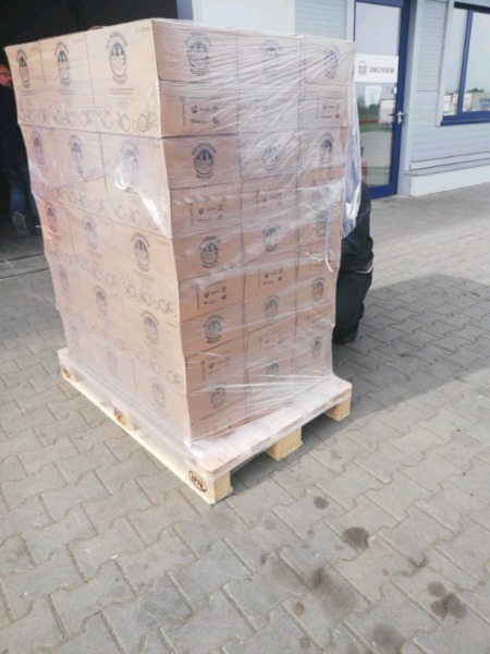 Paleta z kartonami, zabezpieczona folią stretch, na kostce brukowej przed budynkiem z niebieskimi oknami. Częściowo widoczna osoba w ciemnych spodniach.