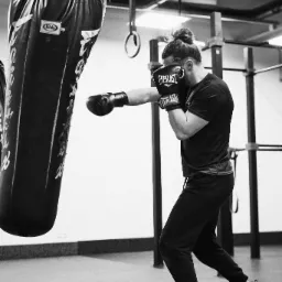 Czarno-białe zdjęcie mężczyzny w czarnych rękawicach bokserskich Everlast uderzającego w wiszący, czarny worek treningowy Fairtex w siłowni.