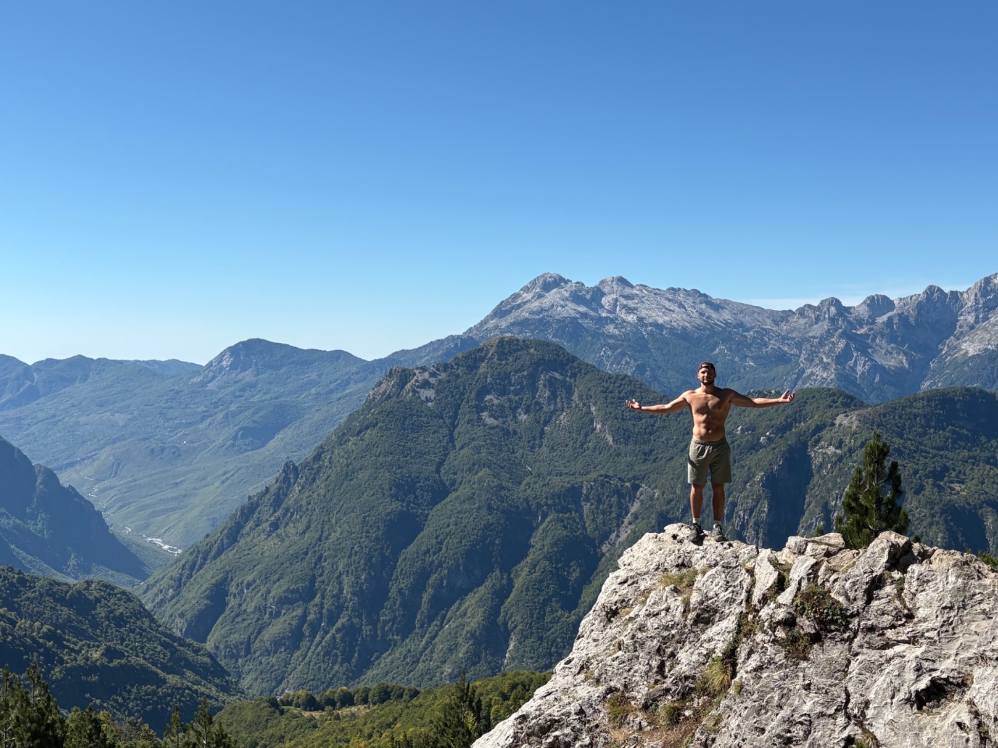 Mężczyzna na szczycie skalistego wzniesienia z rozłożonymi ramionami, w tle rozległy górski krajobraz pod błękitnym niebem. Widok z perspektywy żabiej.
