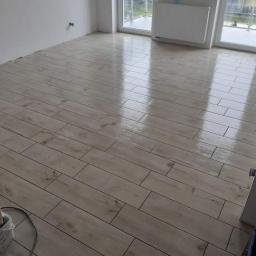 Perfecta - wykończenia wnętrz - Ułożona podłoga z płytek imitujących drewno w jasnym odcieniu, widoczne fugi, błyszcząca powierzchnia odbija światło z okna, w rogu wiaderko z akcesoriami do układania.