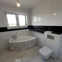 Perfecta - wykończenia wnętrz - Wykończona łazienka z białymi płytkami imitującymi marmur na podłodze i ścianach, z czarnym pasem płytek dekoracyjnych na ścianie, widoczna wanna obudowana mozaiką, wisząca toaleta i okno...