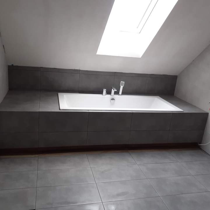 Nowoczesna łazienka na poddaszu z białą wanną obudowaną ciemnoszarą płytką, szara płytka podłogowa i okno dachowe w skosie.