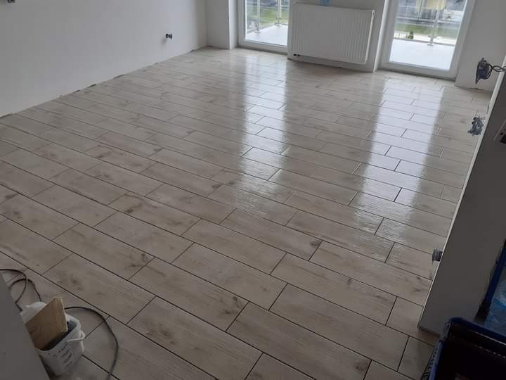 Ułożona podłoga z płytek imitujących drewno w jasnym odcieniu, widoczne fugi, błyszcząca powierzchnia odbija światło z okna, w rogu wiaderko z akcesoriami do układania.