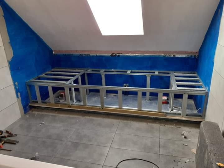 Łazienka na poddaszu w trakcie tworzenia zabudowy z profili metalowych pod wannę lub brodzik, ściany zabezpieczone folią w płynie, widoczne narzędzia na podłodze z płytek.