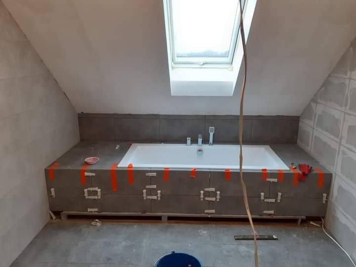 Łazienka na poddaszu w trakcie remontu: wanna obudowana szarymi płytkami, zabezpieczona taśmą, widoczne elementy instalacji i narzędzia, okno dachowe.