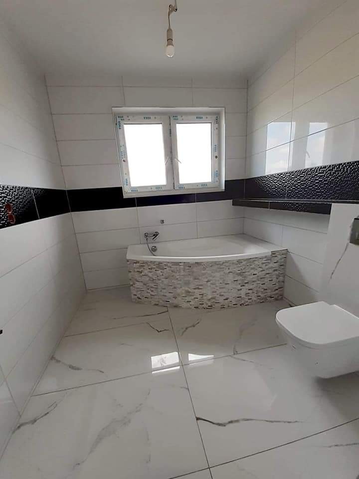 Nowoczesna łazienka z biało-czarną glazurą, marmurową podłogą i obudową wanny z mozaiki, widok na okno z białymi ramami.